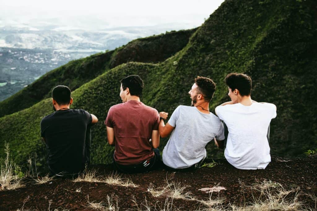 Four young men seated on a lush green mountain top, enjoying the panoramic view below, symbolizing camaraderie, adventure, and connection with nature.