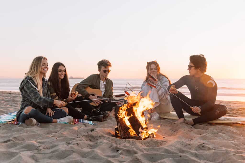 A group of young people gathered on the beach around a bonfire at sunset. The image captures the warmth of friendship and the beauty of a beach sunset, creating a relaxed and joyful atmosphere. The silhouettes of the group against the vibrant sky evoke a sense of camaraderie and shared moments.