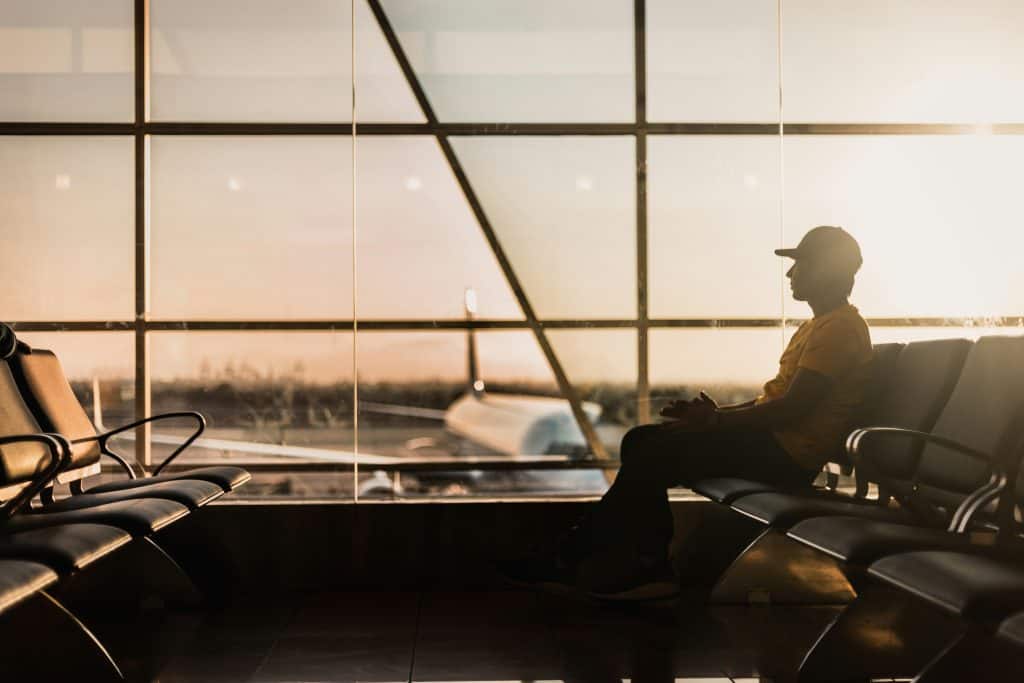 Man sitting at an airport gate, looking out the window at planes on the tarmac, with a carry-on bag by his side.