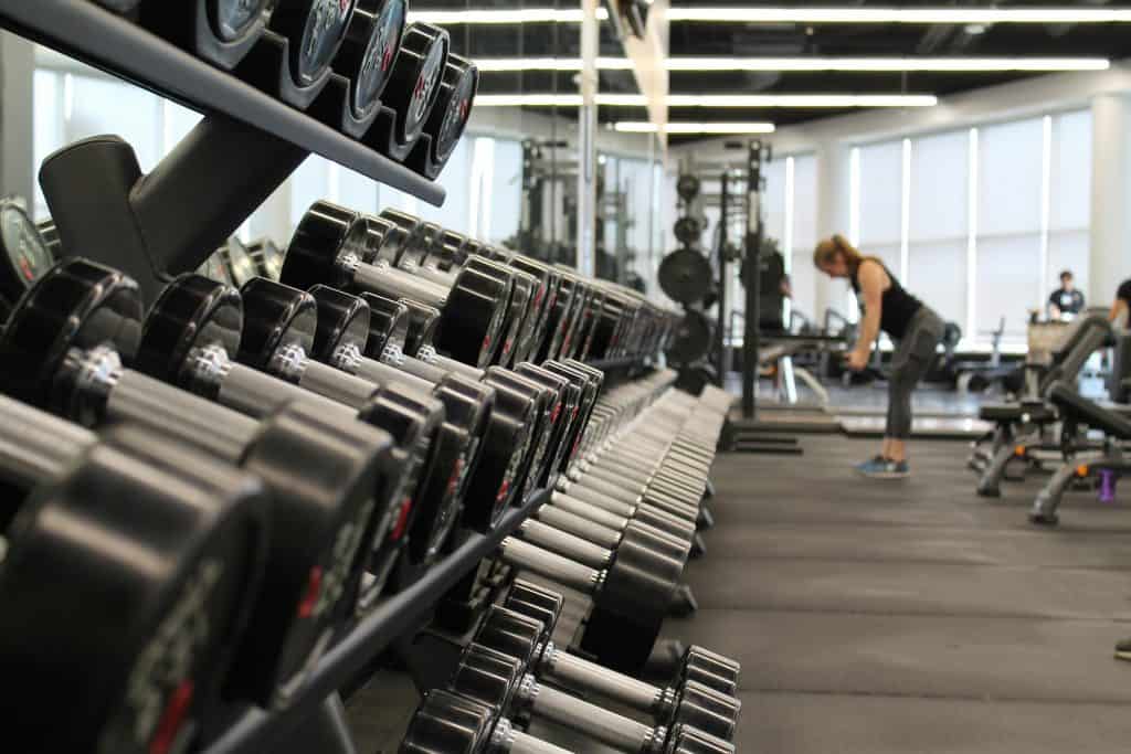 A rack of dumbbells in a gym with a woman exercising in the background, capturing the focus on strength training and dedication to fitness.