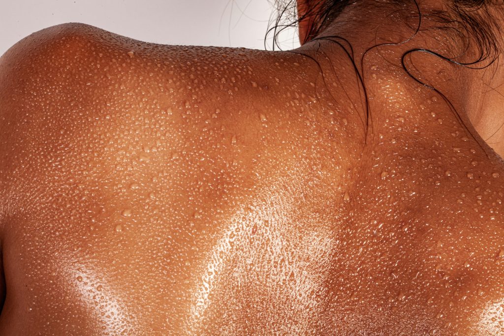 Close-up of a woman's back covered in beads of sweat, highlighting her toned skin glistening under soft light, emphasizing the effects of a detoxifying workout or sauna session.