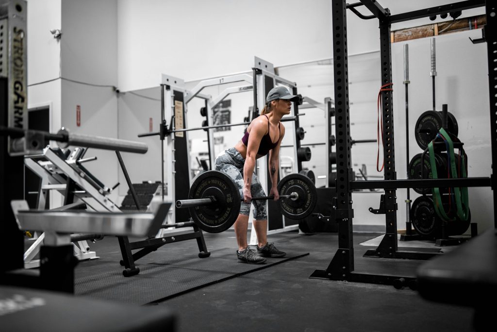 A woman performing a barbell deadlift in a gym setting, surrounded by weightlifting equipment, wearing athletic gear and a cap, focusing on strength training.