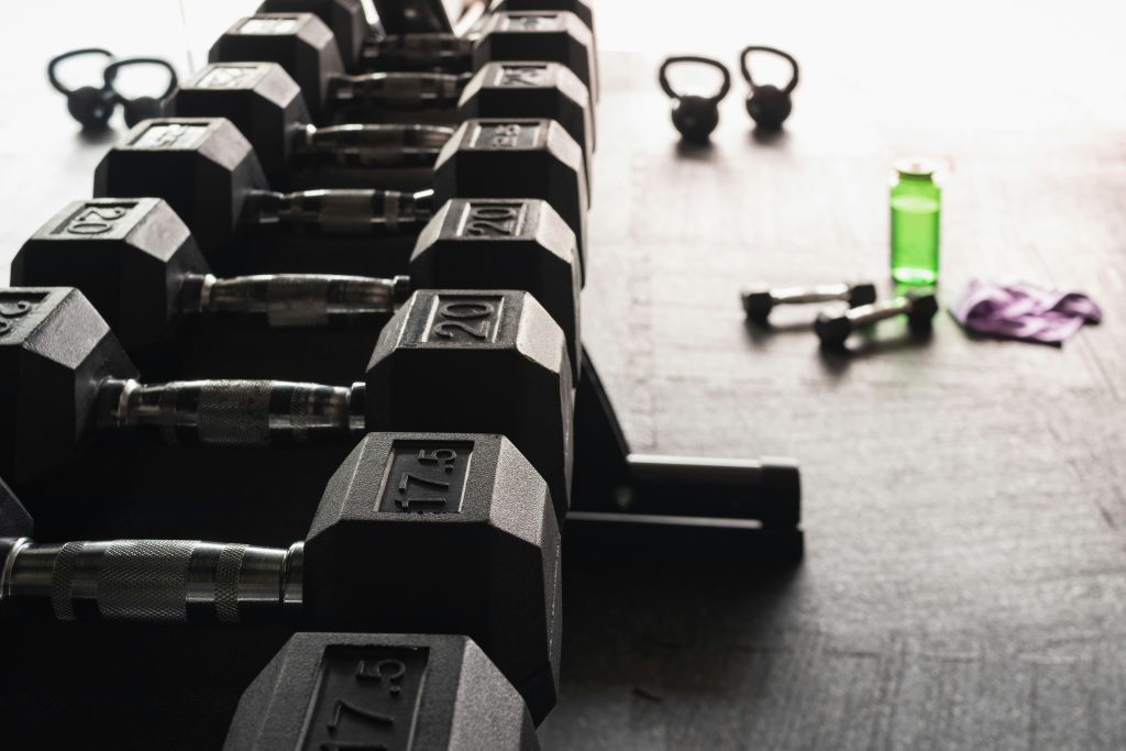 A rack of black dumbbells neatly arranged, with kettlebells, a green water bottle, and a purple towel in the background on a gym floor.