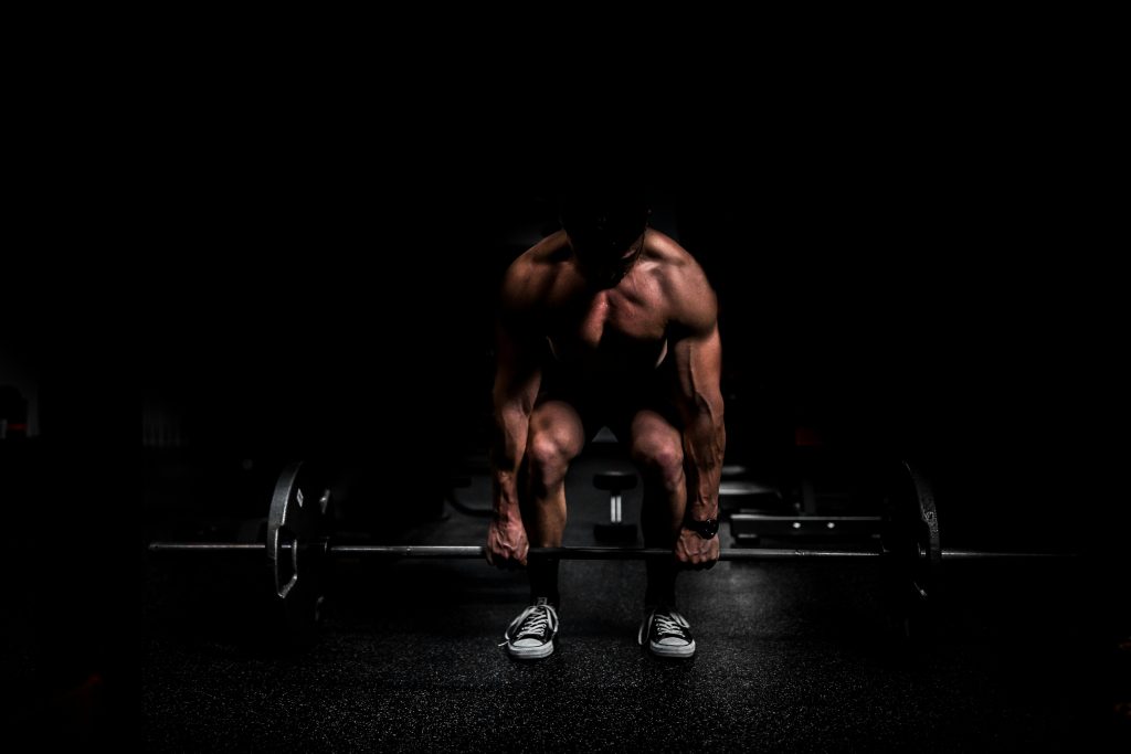 A muscular man performing a deadlift in a dimly lit gym. The focus is on his tense posture and the barbell he is lifting, highlighting strength training and intensity in progressive overload exercises.