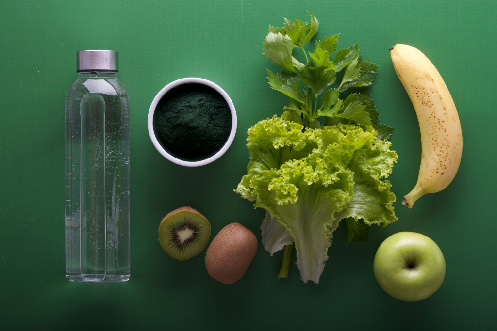 Flat lay of a water bottle, spirulina powder, kiwi, lettuce, banana, and apple representing electrolyte-rich foods and hydration