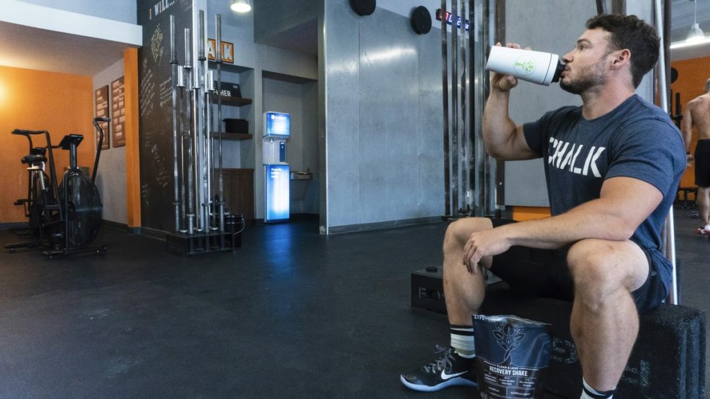 Athlete resting on a gym floor while drinking a post-workout recovery shake