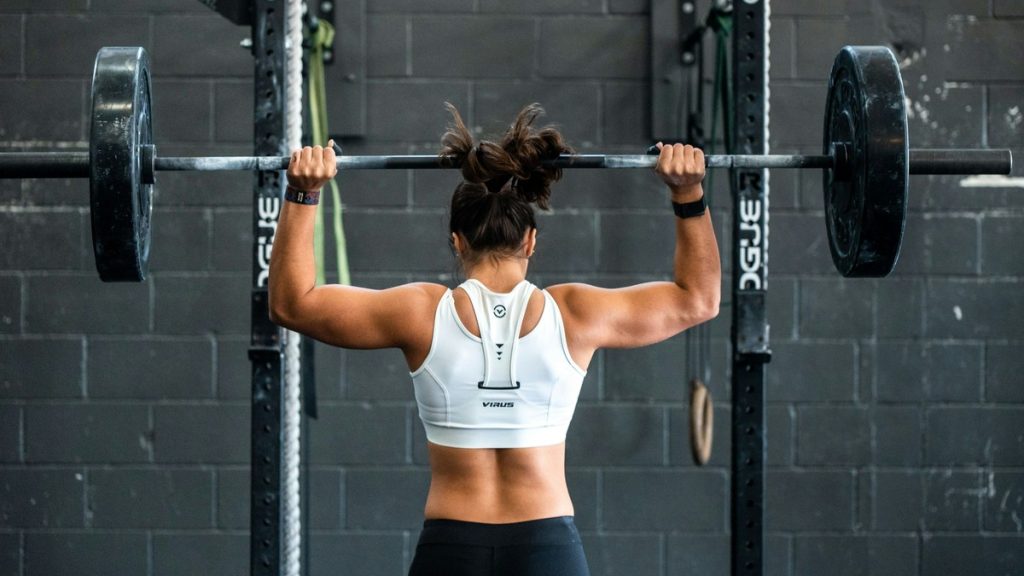 Woman performing an overhead barbell press from behind in a gym, focused and strong