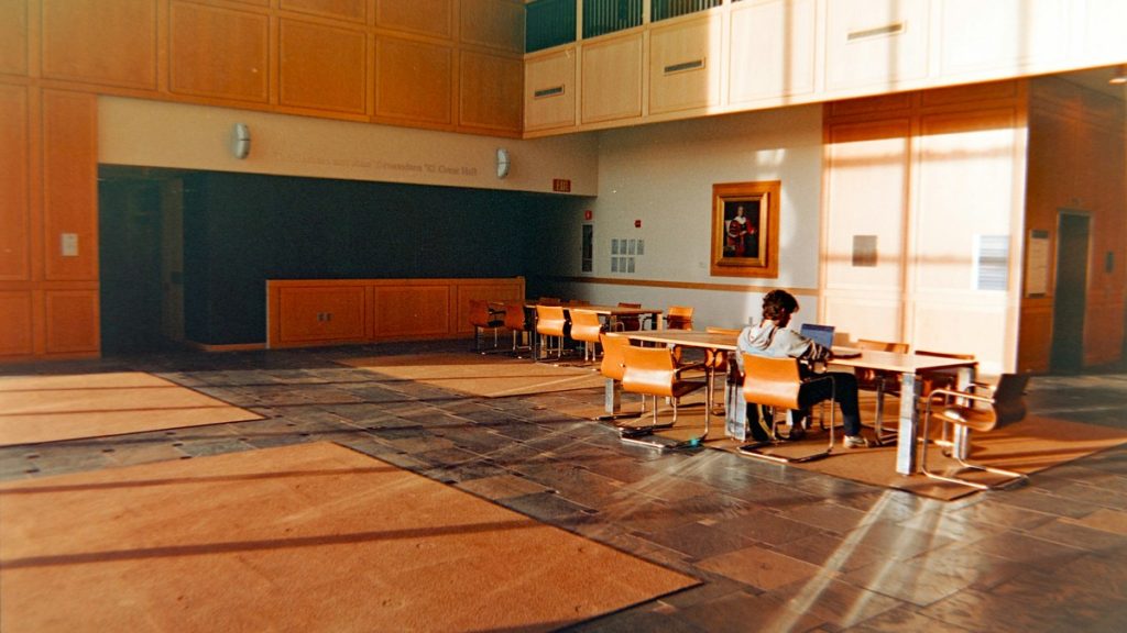 College student studying alone with a laptop at a table in a sunlit campus atrium
