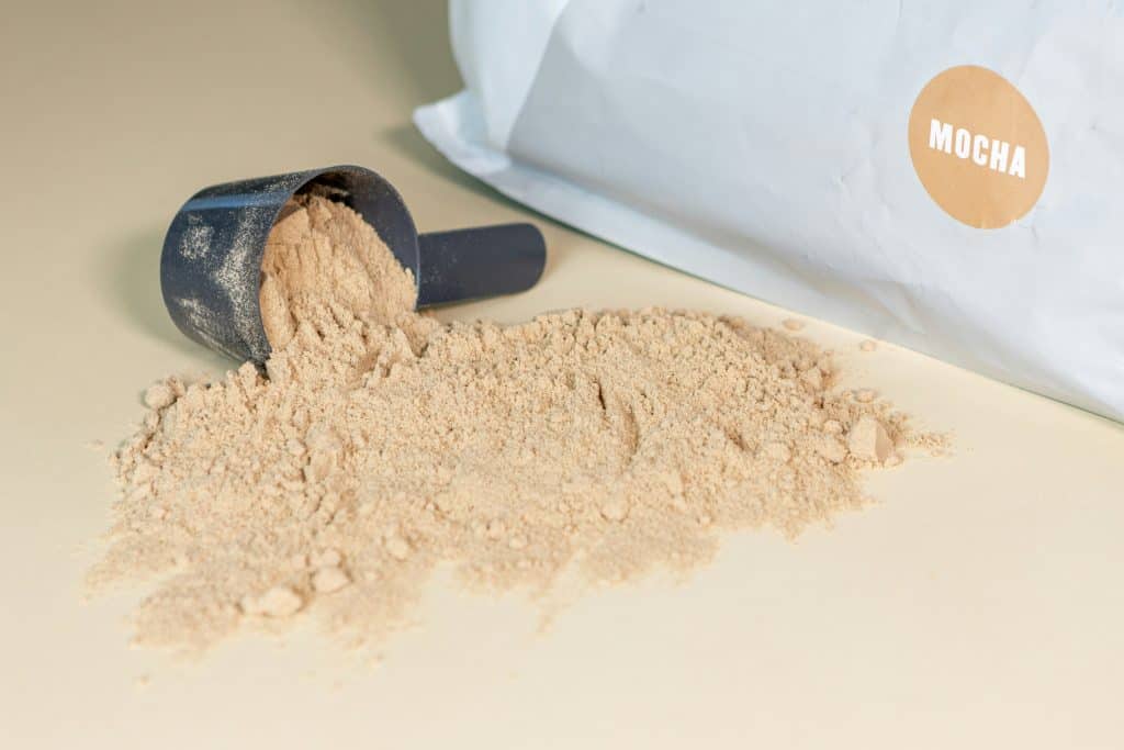 Container of protein powder beside a full measuring scoop on a light surface