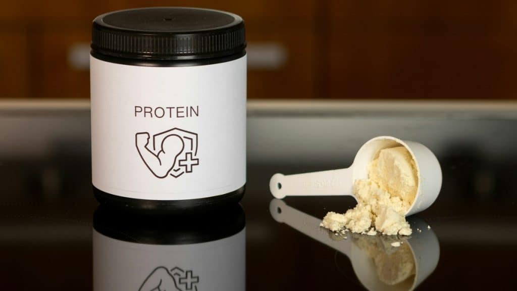 Container of protein powder beside a full measuring scoop on a dark surface